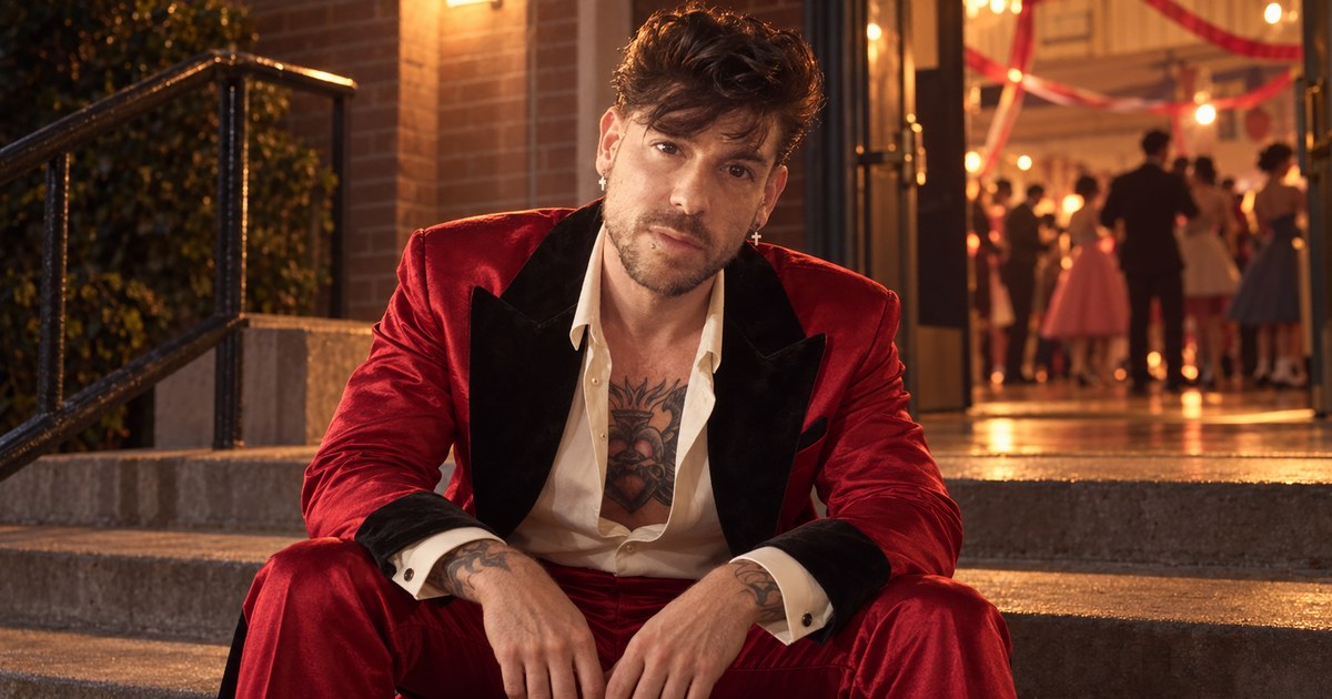 Mike Saint Louis con traje de terciopelo rojo y solapas negras, sentado en las escaleras de piedra de un edificio de ladrillo de noche, con un baile iluminado al fondo.