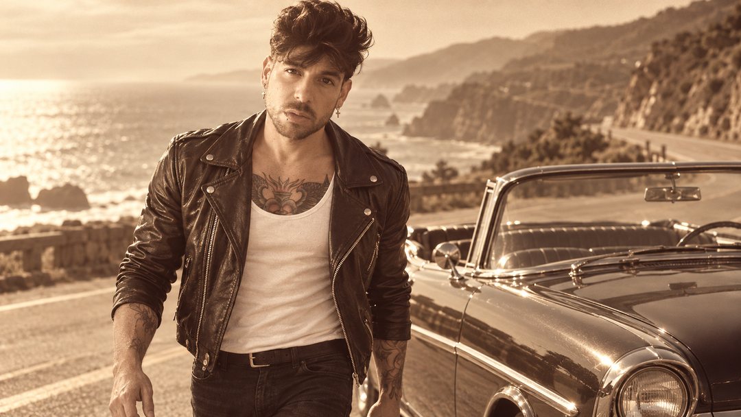 Mike Sain't Louis con chaqueta de cuero y camiseta blanca junto a un descapotable clásico en una carretera de costa — foto retro PLANETA GAYFOLK.