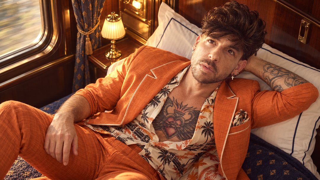 Mike Sain't Louis con traje naranja y camisa estampada recostado en la litera de un coche cama antiguo — retrato Tupé Grease PLANETA GAYFOLK.