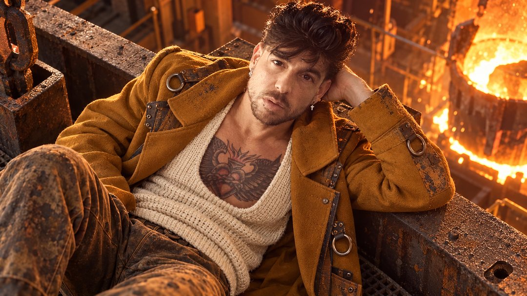 Mike Sain't Louis con chaqueta mostaza y camiseta de punto recostado junto a un horno encendido en una fundición — retrato Tupé Grease PLANETA GAYFOLK.
