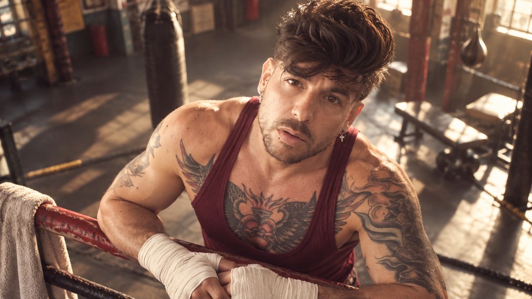 Mike Sain't Louis con camiseta de tirantes burdeos y vendas en las manos apoyado en las cuerdas de un ring en un gimnasio antiguo — retrato Tupé Grease PLANETA GAYFOLK.