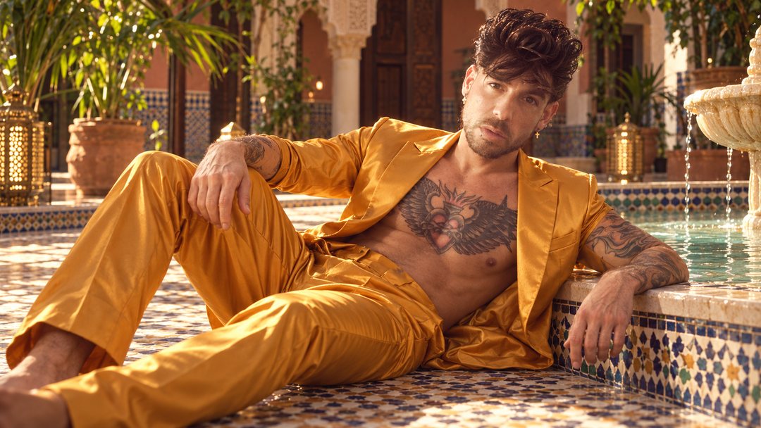Mike Sain't Louis con traje de satén dorado recostado junto a una piscina de azulejos en un patio andaluz — retrato Tupé Grease PLANETA GAYFOLK.