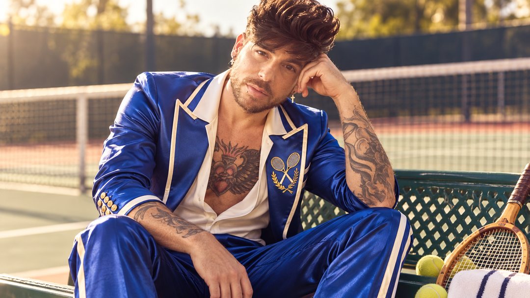 Mike Sain't Louis con chándal de satén azul con ribetes blancos sentado en un banco junto a una pista de tenis — retrato Tupé Grease PLANETA GAYFOLK.
