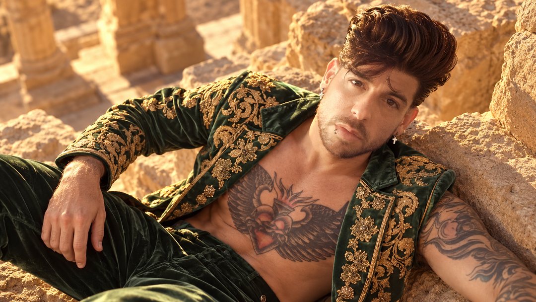 Mike Sain't Louis con traje de terciopelo verde bordado en oro recostado entre rocas de arenisca al sol — retrato Tupé Grease PLANETA GAYFOLK.