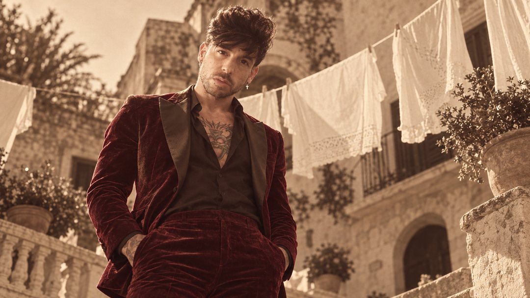 Mike Sain't Louis con traje de terciopelo burdeos, único toque de color, en una calle de pueblo con ropa tendida — foto retro PLANETA GAYFOLK.