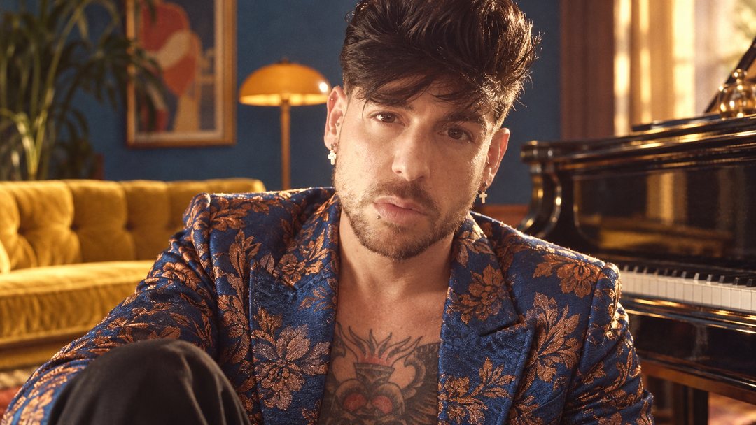 Mike Sain't Louis con chaqueta de brocado azul y dorado junto a un piano en un salón con sofá amarillo — foto retro PLANETA GAYFOLK.