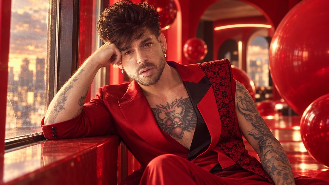 Mike Sain't Louis con traje de raso rojo recostado junto a una ventana en un salón rojo con esferas rojas y vistas a la ciudad — retrato Tupé Grease PLANETA GAYFOLK.
