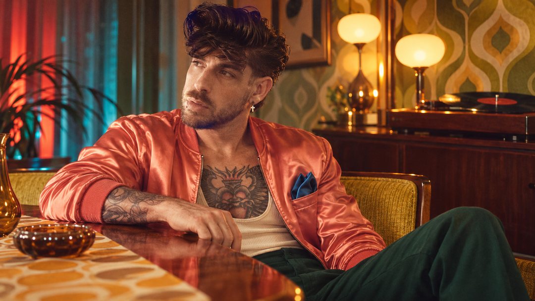 Mike Sain't Louis con cazadora satinada coral y pantalón verde en un salón de estilo setentero — foto retro PLANETA GAYFOLK.