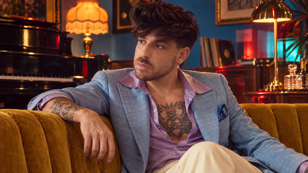 Mike Sain't Louis con americana azul claro y camisa lila sentado en un sofá de terciopelo mostaza — foto retro PLANETA GAYFOLK.
