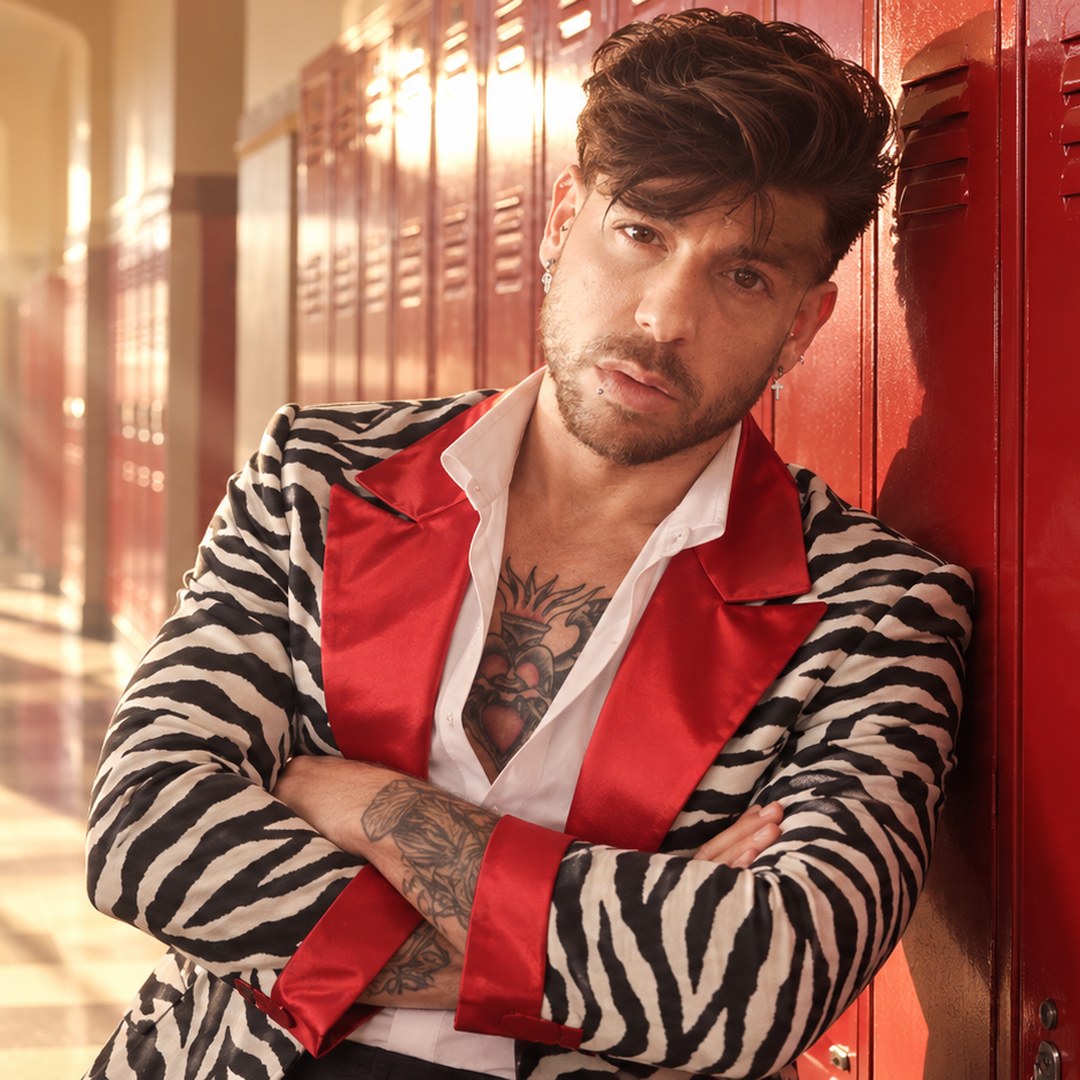 Mike Sain't Louis con chaqueta de cebra y solapas rojas apoyado en unas taquillas rojas de un pasillo escolar — foto PLANETA GAYFOLK.
