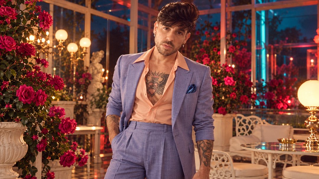 Mike Sain't Louis con traje lavanda en una terraza con rosas y mesas de hierro blanco de noche — foto retro PLANETA GAYFOLK.