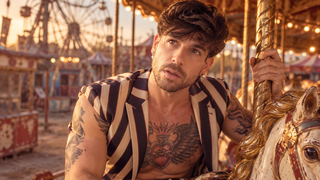 Mike Sain't Louis con chaleco de rayas blancas y negras junto a un caballito de tiovivo con una noria al fondo — retrato Tupé Grease PLANETA GAYFOLK.